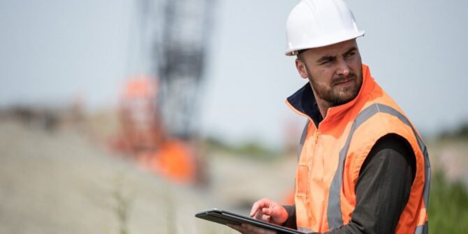 A photograph of a man in a bright orange safety vest and white hard hat using an iPad