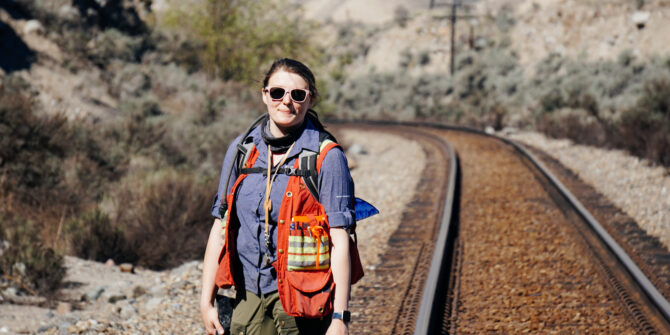 A person in a surveyor's vest and sunglasses stands on a railroad track in a desert, holding a geological rock hammer