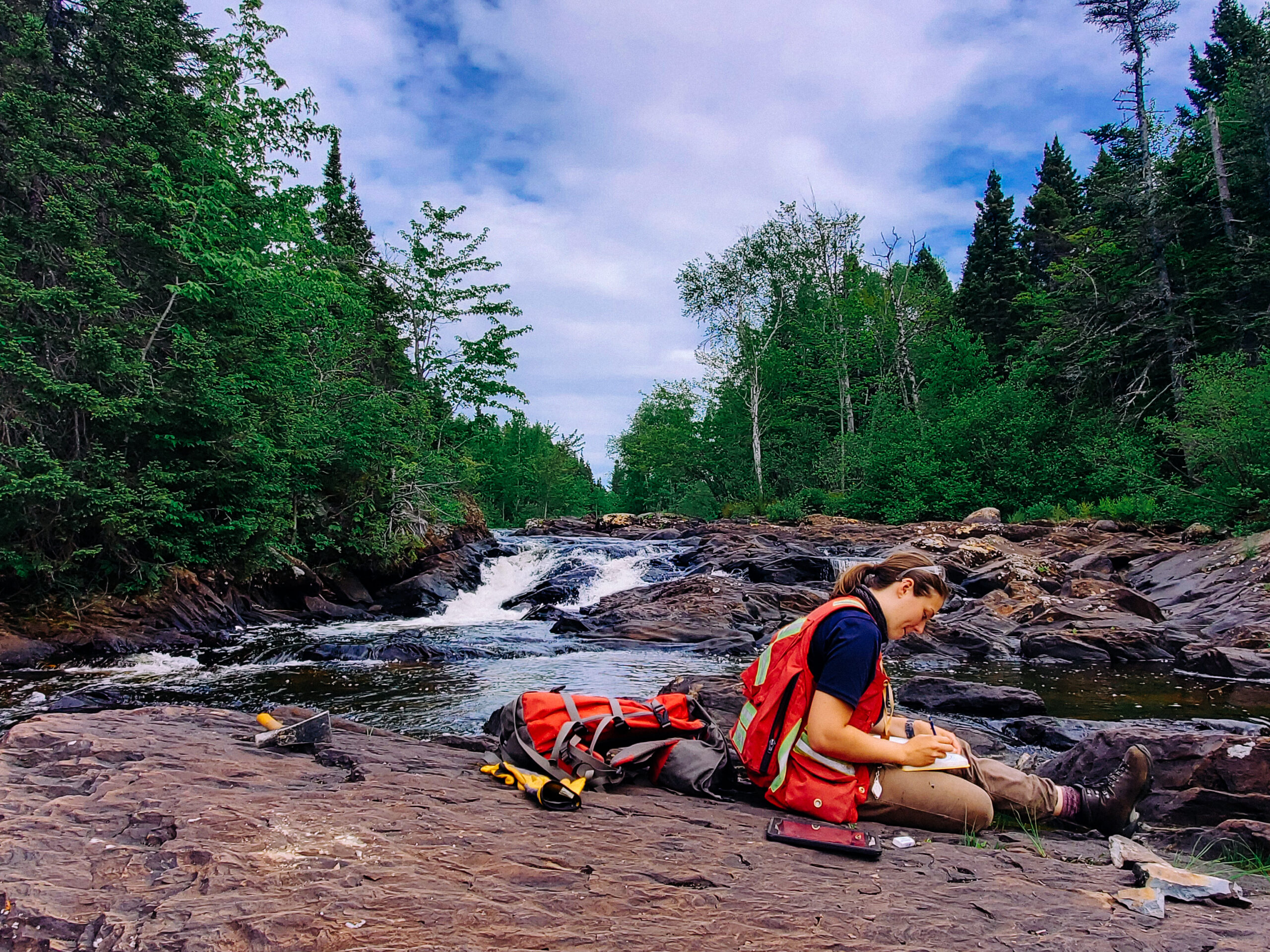 A geologist wearing a high-visibility vest sits on a rocky riverbank, recording field notes near a cascading waterfall. Surrounded by a dense forest, she works alongside her geological hammer and field pack.