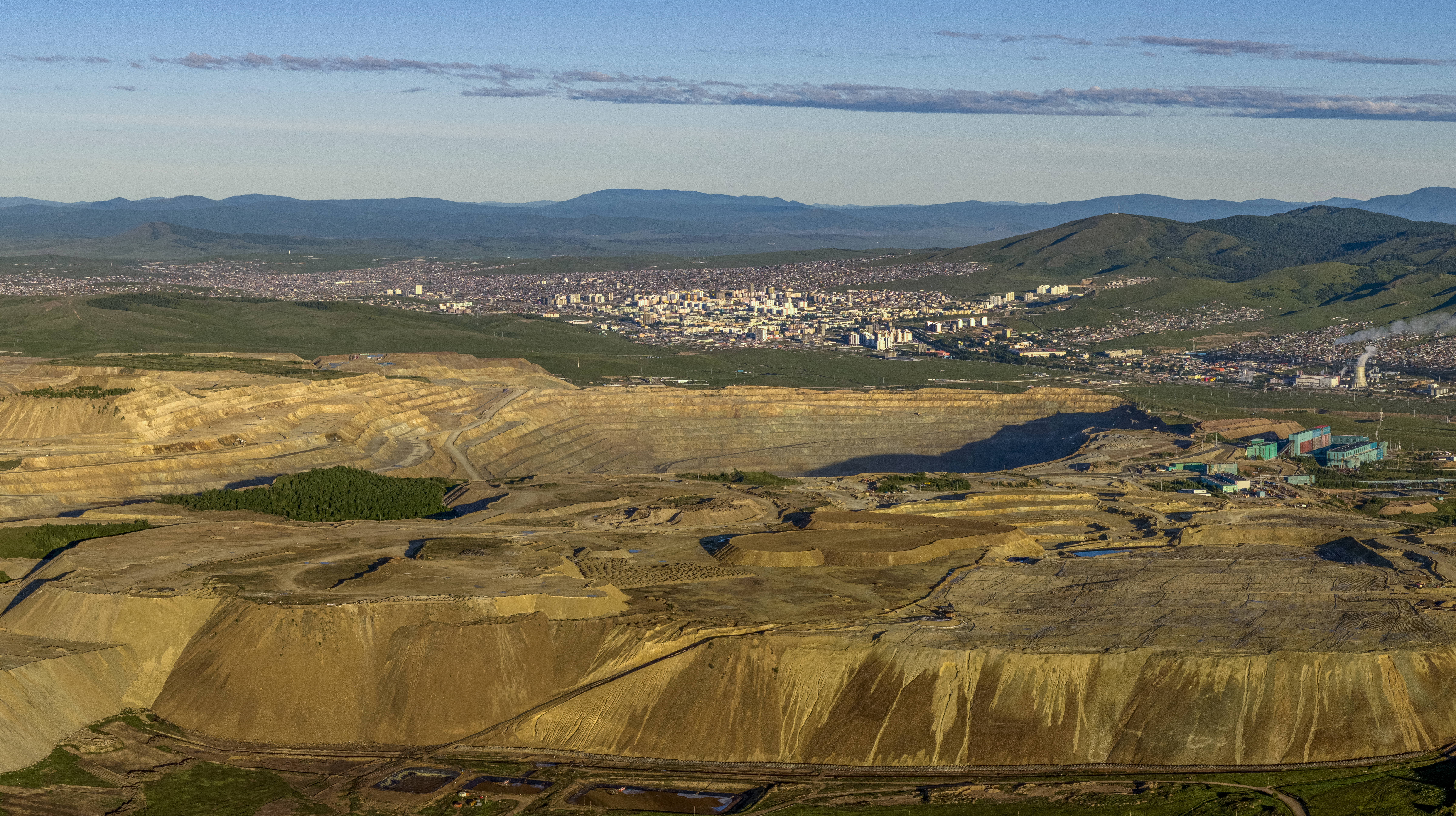Aerial view of an open-pit mine with terraced walls in the foreground and a large city nestled against rolling hills in the background.