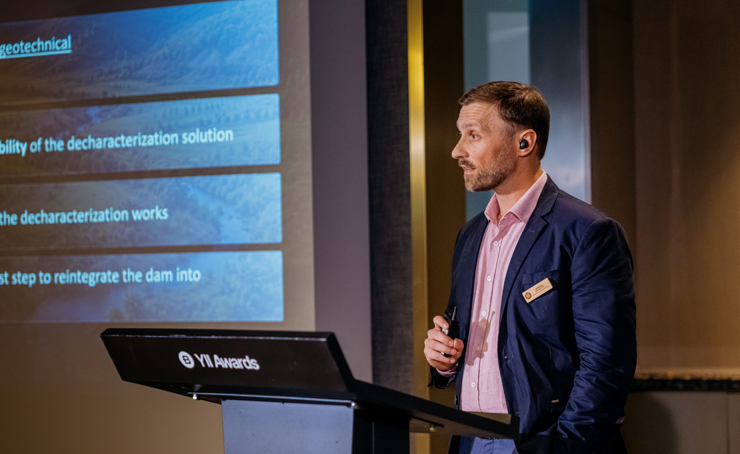 Marcello Miqueletto from Intertechne Consultores S.A during his finalist presentation stands at a lectern beside a projected slide with blue text panels about geotechnical work and dam decharacterization.