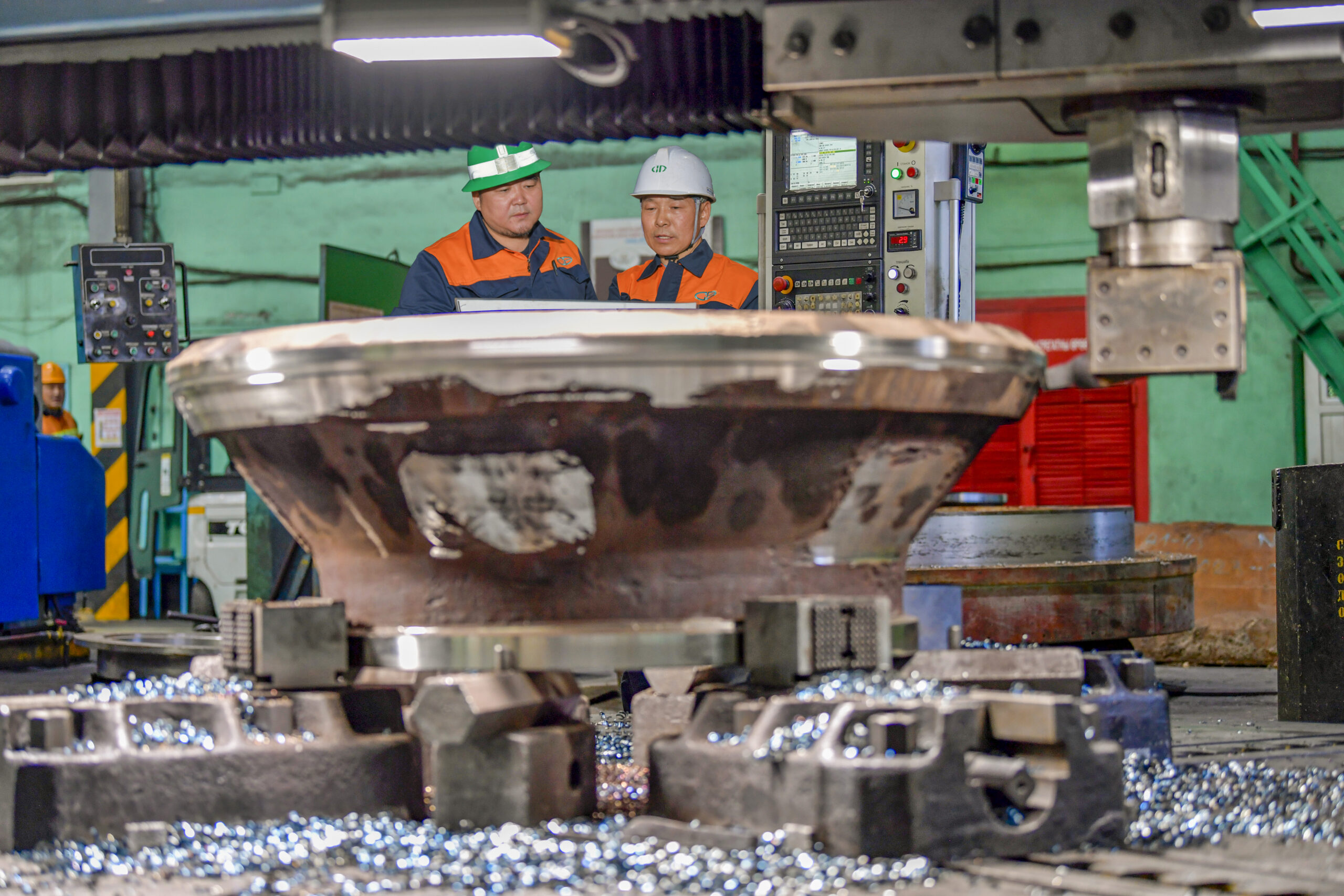 Two technicians in hard hats and safety gear inspecting a large metal component being machined on a lathe in an industrial workshop