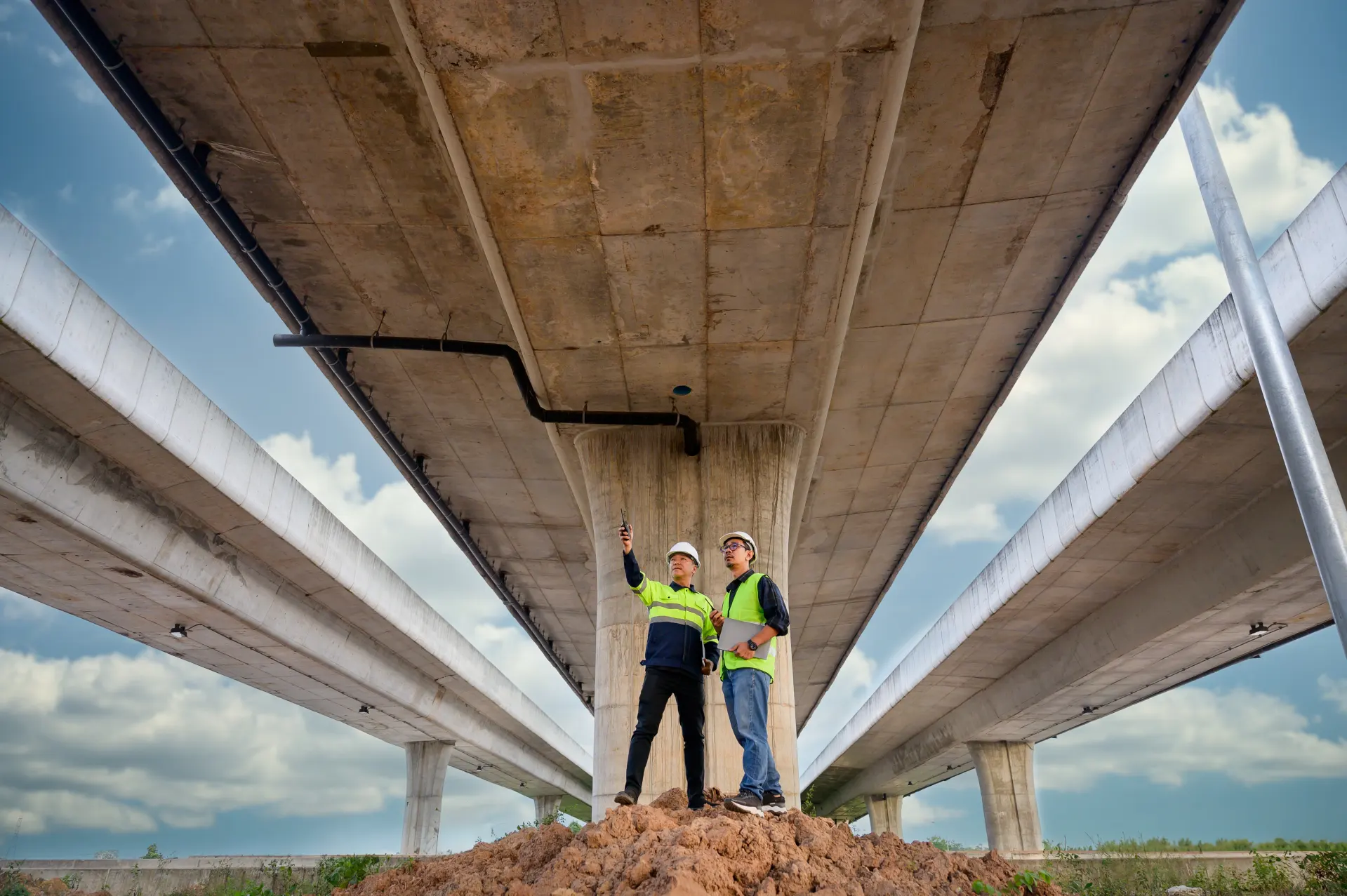 Two engineers in hard hats and safety vests inspecting the underside of a large concrete highway overpass.