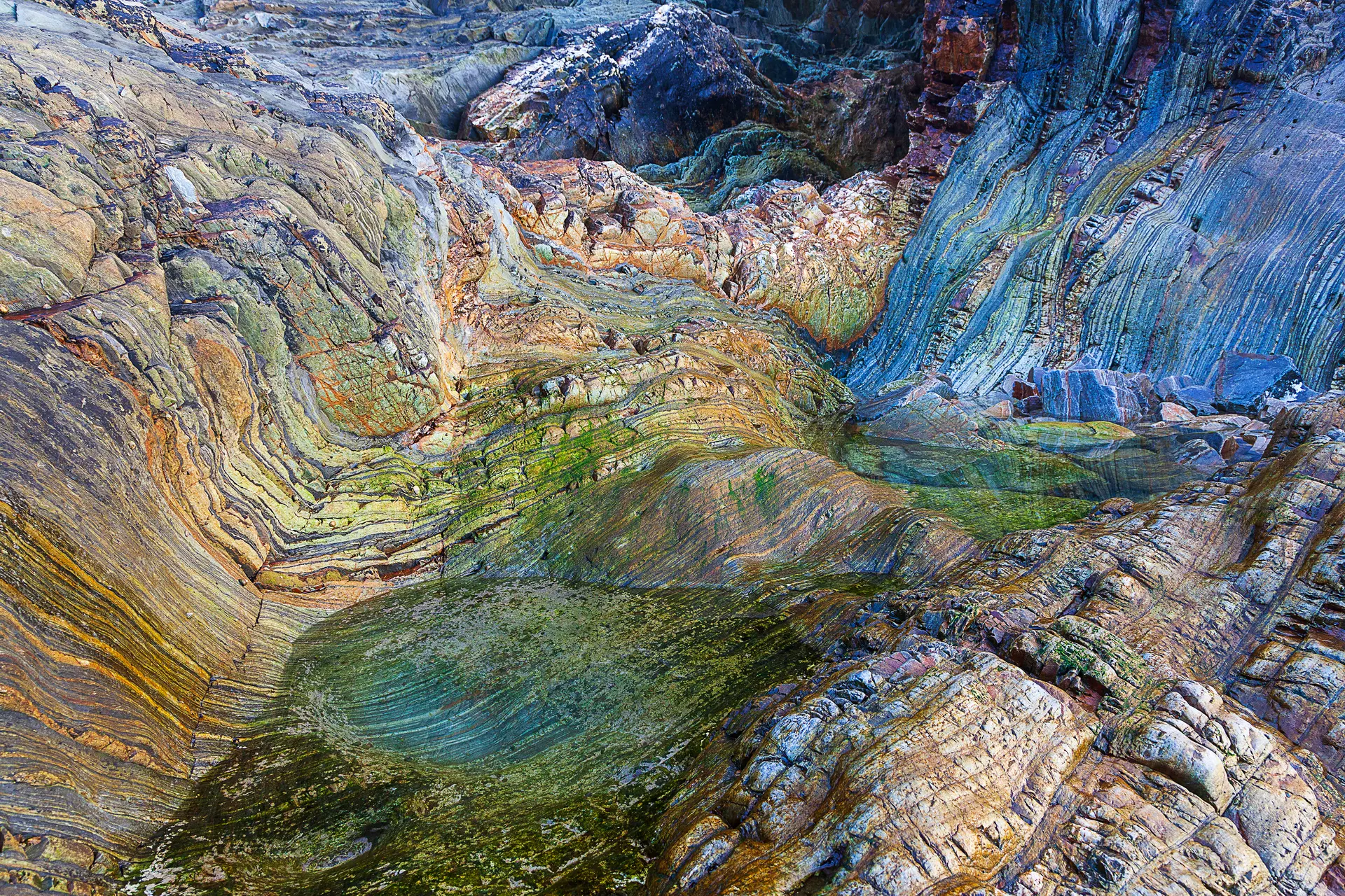 A photo of rock formations showing distinct, colourful layers and bands formed over long periods, indicative of metamorphic or sedimentary processes. The texture shows extreme terrain, likely shaped by erosion from wind and water.