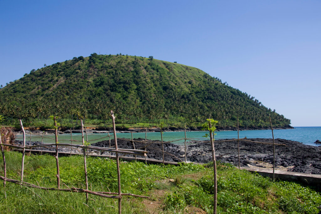A lush, green tropical hill rises above a turquoise bay. In the foreground, a rustic wooden stick fence borders a grassy area next to dark, volcanic rocks along the shoreline under a clear blue sky.
