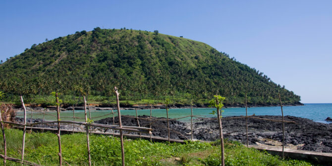 A lush, green tropical hill rises above a turquoise bay. In the foreground, a rustic wooden stick fence borders a grassy area next to dark, volcanic rocks along the shoreline under a clear blue sky.