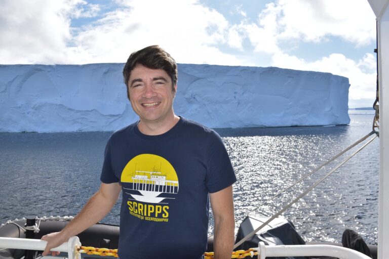 Polar Scientist Dr Jamin Greenbaum stands on a boat with the ocean and an iceberg in the background.