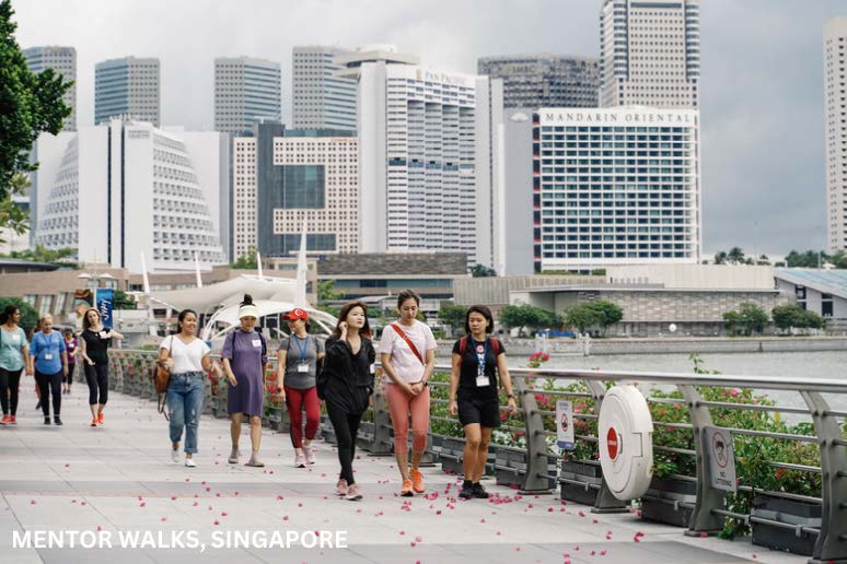 A photograph of a group women walking on a path with a city in the background captioned Mentor Walks, Singapore