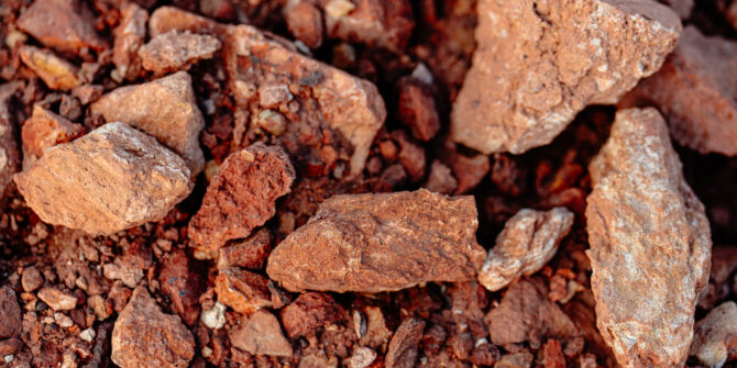 A top-down, close-up view of rough, reddish-brown iron ore rocks and soil of varying sizes and textures.