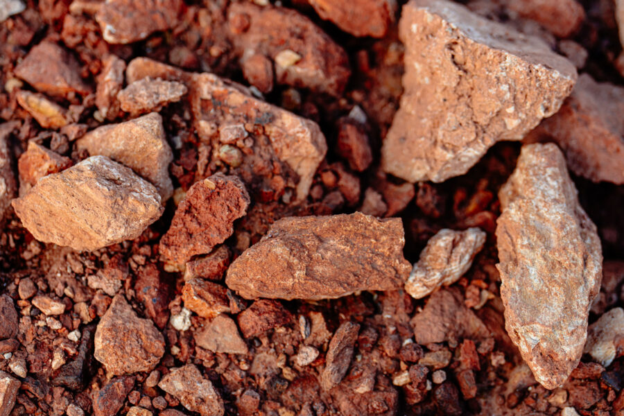 A top-down, close-up view of rough, reddish-brown iron ore rocks and soil of varying sizes and textures.