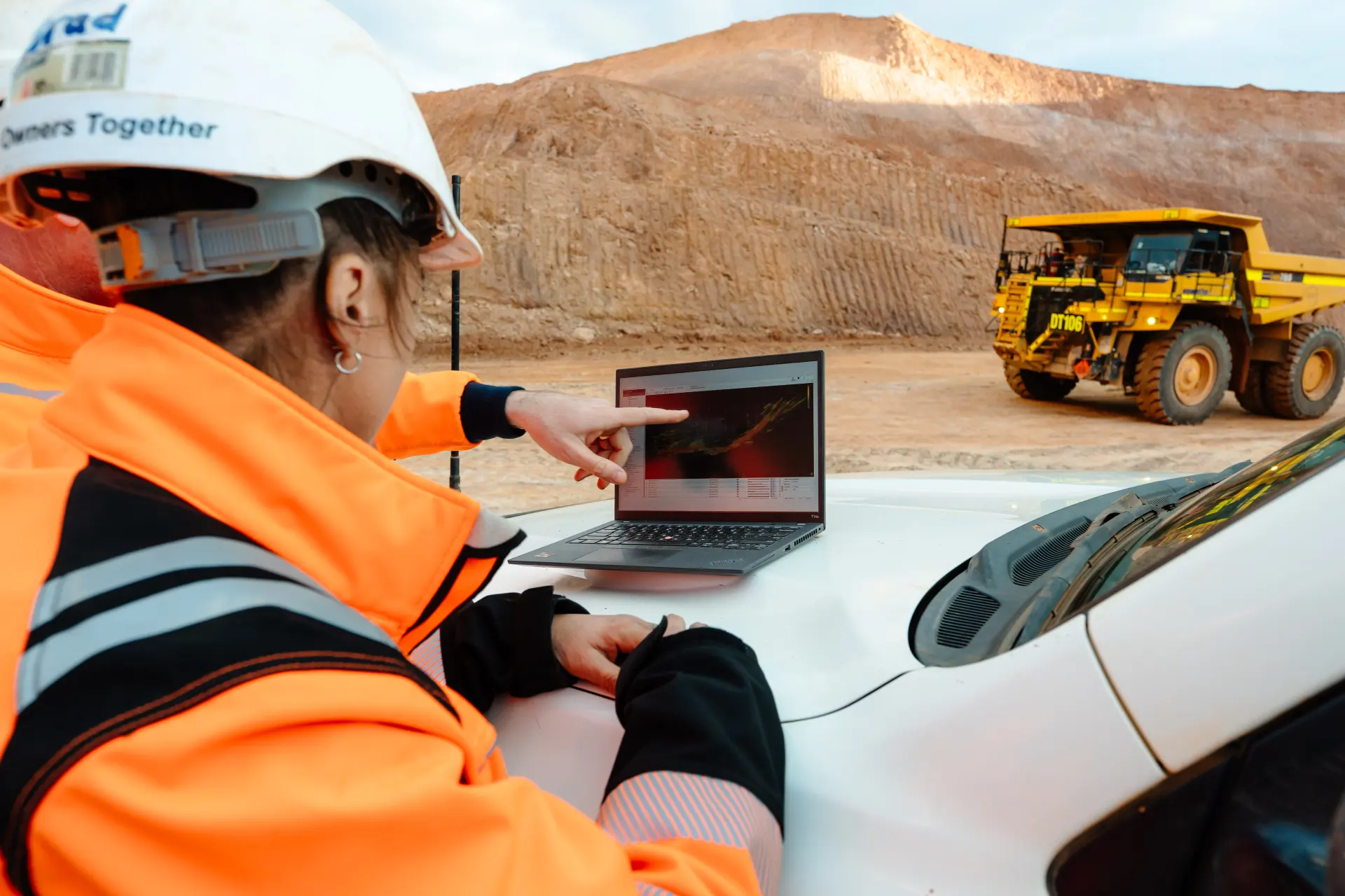 At Mt Ida in Western Australia, part of Aurenne Group Mining’s open‑pit gold mining operation, a worker in high‑visibility safety clothing reviews geological or site data on a laptop placed on a vehicle, with haul trucks and pit walls visible in the background. The image reflects on‑site operational monitoring and decision‑making during active mining activities. Aurenne Photoshoot 2025.