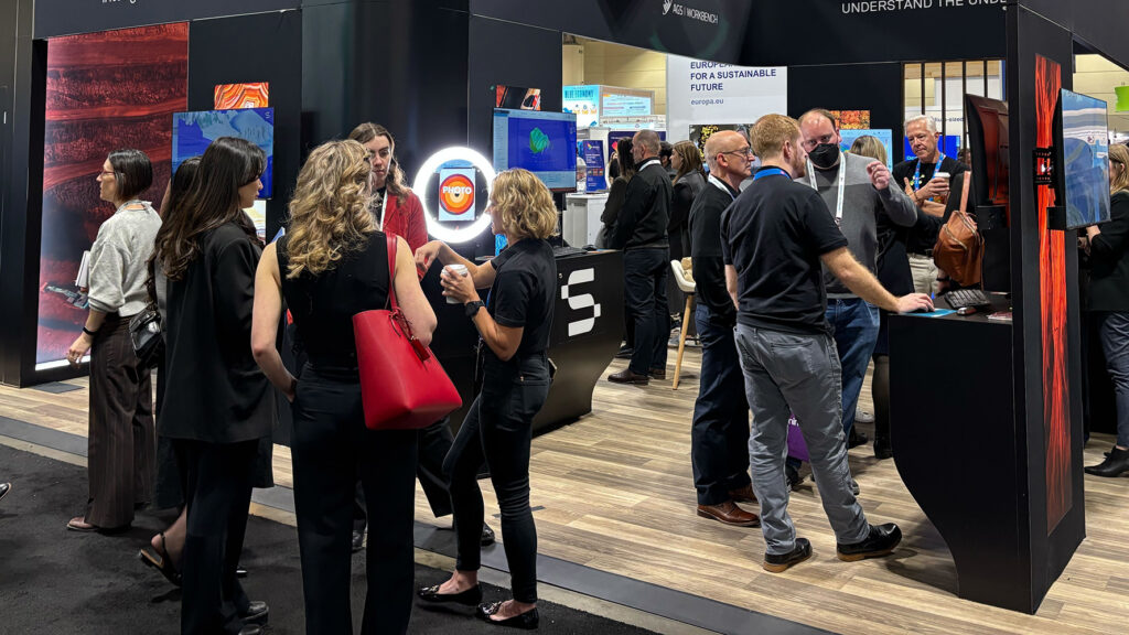 Scene from the PDAC mineral exploration and mining convention showing a crowd of industry professionals. A banner for the PDAC event in Toronto is prominently displayed in the background.