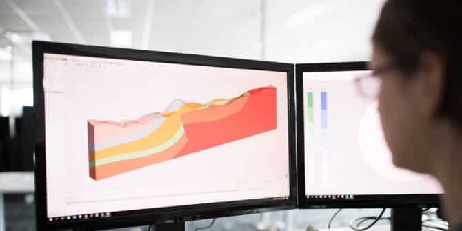 A photograph of a woman looking at 2 screens with 3D geological models visible on them