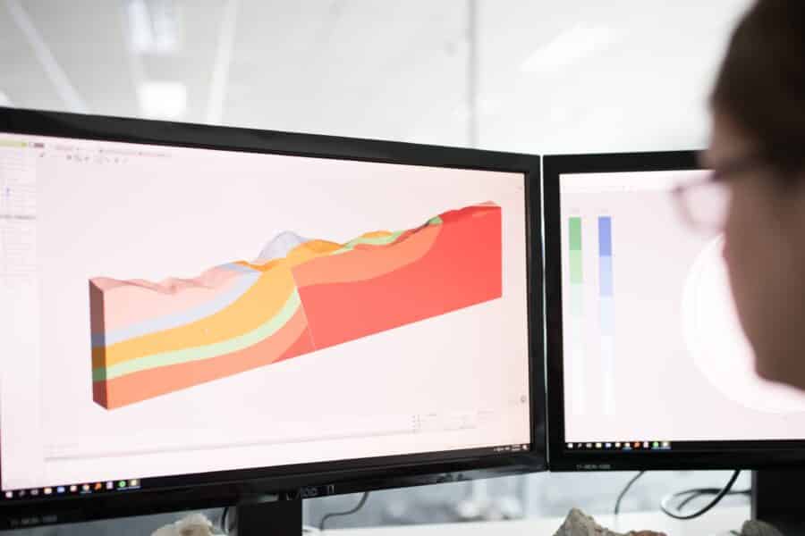 A photograph of a woman looking at 2 screens with 3D geological models visible on them