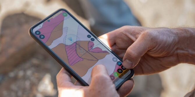 Close-up of a person's hands holding a smartphone displaying a colorful digital topographical map while outdoors