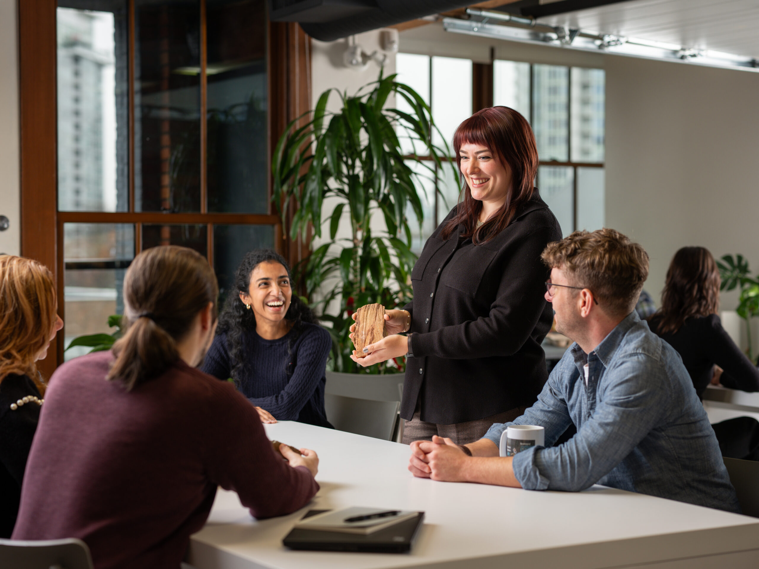 A group of professionals in an office setting, where one woman stands at a table holding a rock sample for her colleagues to see. The team is engaged in a collaborative discussion, likely regarding geological data or resource modeling