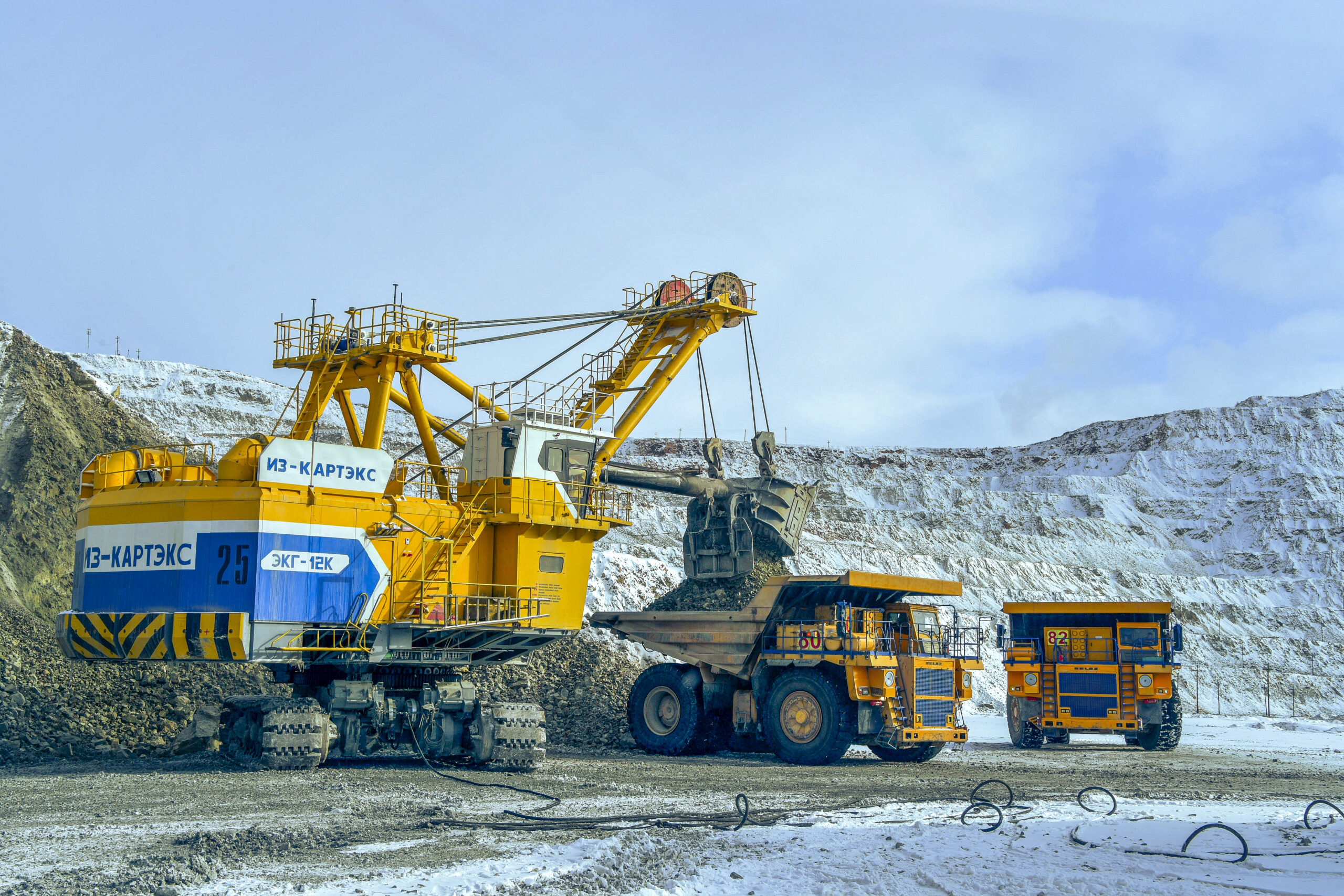 An electric rope shovel loads ore into a haul truck at a snow-dusted open-pit mine, with a second truck waiting nearby
