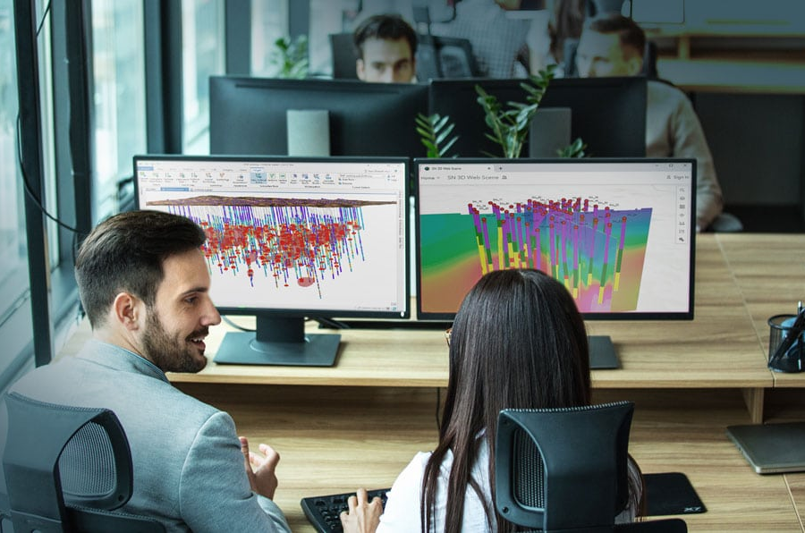 Two colleagues in an office collaborating over dual monitors that are displaying complex 3D models of geological data.