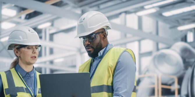 Two Diverse Professional Heavy Industry Engineers Wearing Safety Uniform and Hard Hats Working on Laptop Computer. African American Technician and Female Worker Talking on a Meeting in a Factory.