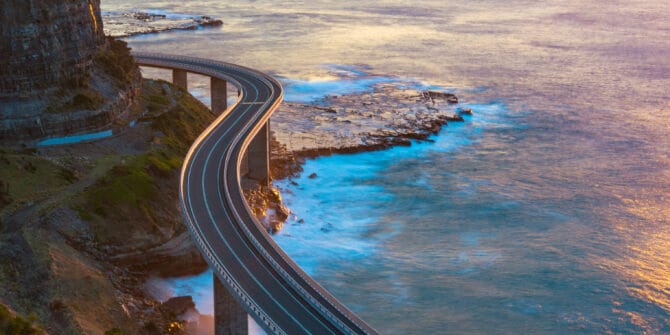 A photograph of a curved road on pillars along a mountainous coastline at sunset