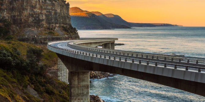 A photograph of a curved road on pillars along a mountainous coastline at sunset